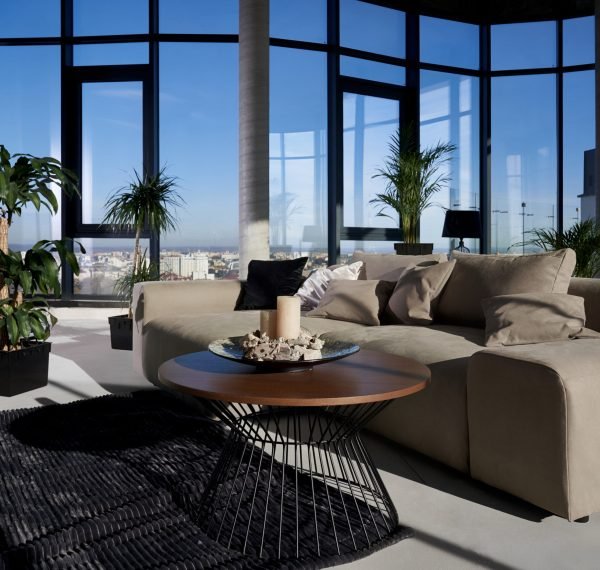 Front view of fashionable interior spacious room with large panoramic window with incredible view on city and cozy grey sofa with pillows and green flowerpots. Concept of modern interior.